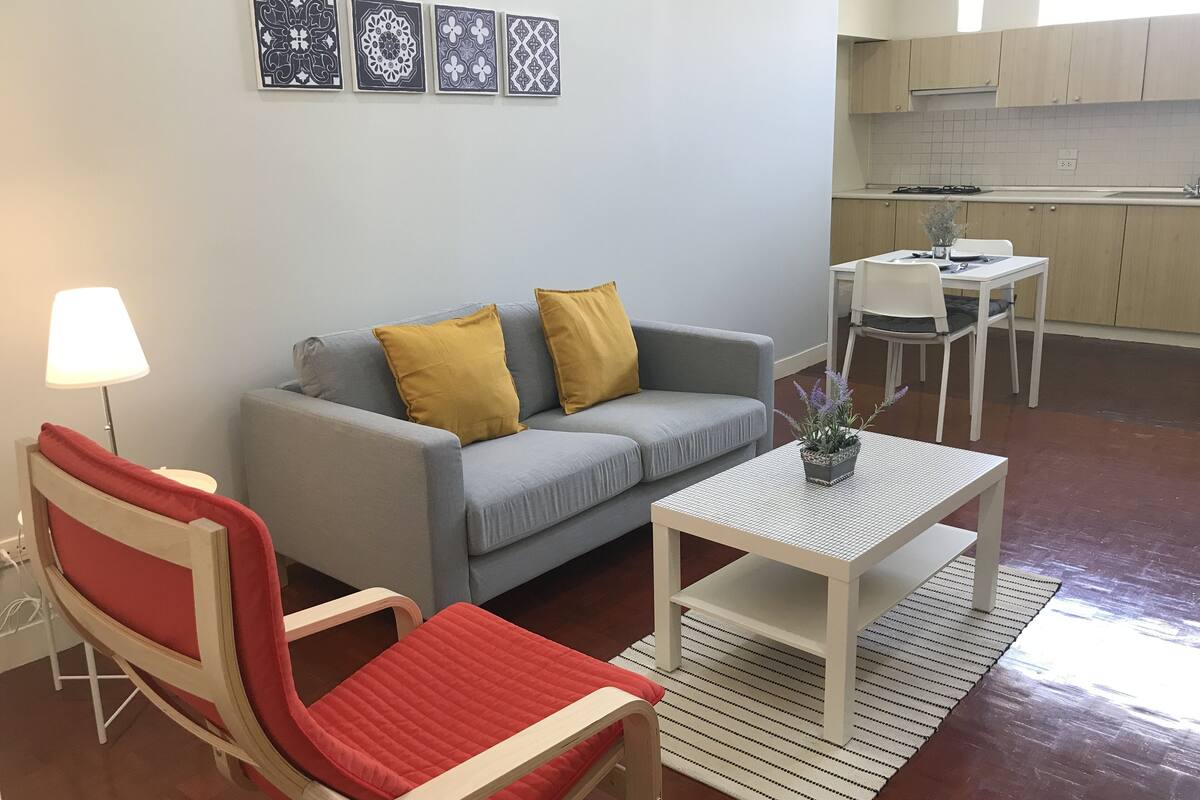 Una sala de estar con un sofá gris, dos cojines amarillos, un sillón rojo, una mesa blanca y una pequeña planta en maceta.
