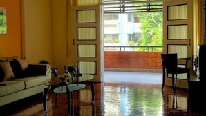 A room with a sofa, a small round table, and a chair. There is a large window with a view of a brick wall and greenery outside. - PT Court (Bangkok)