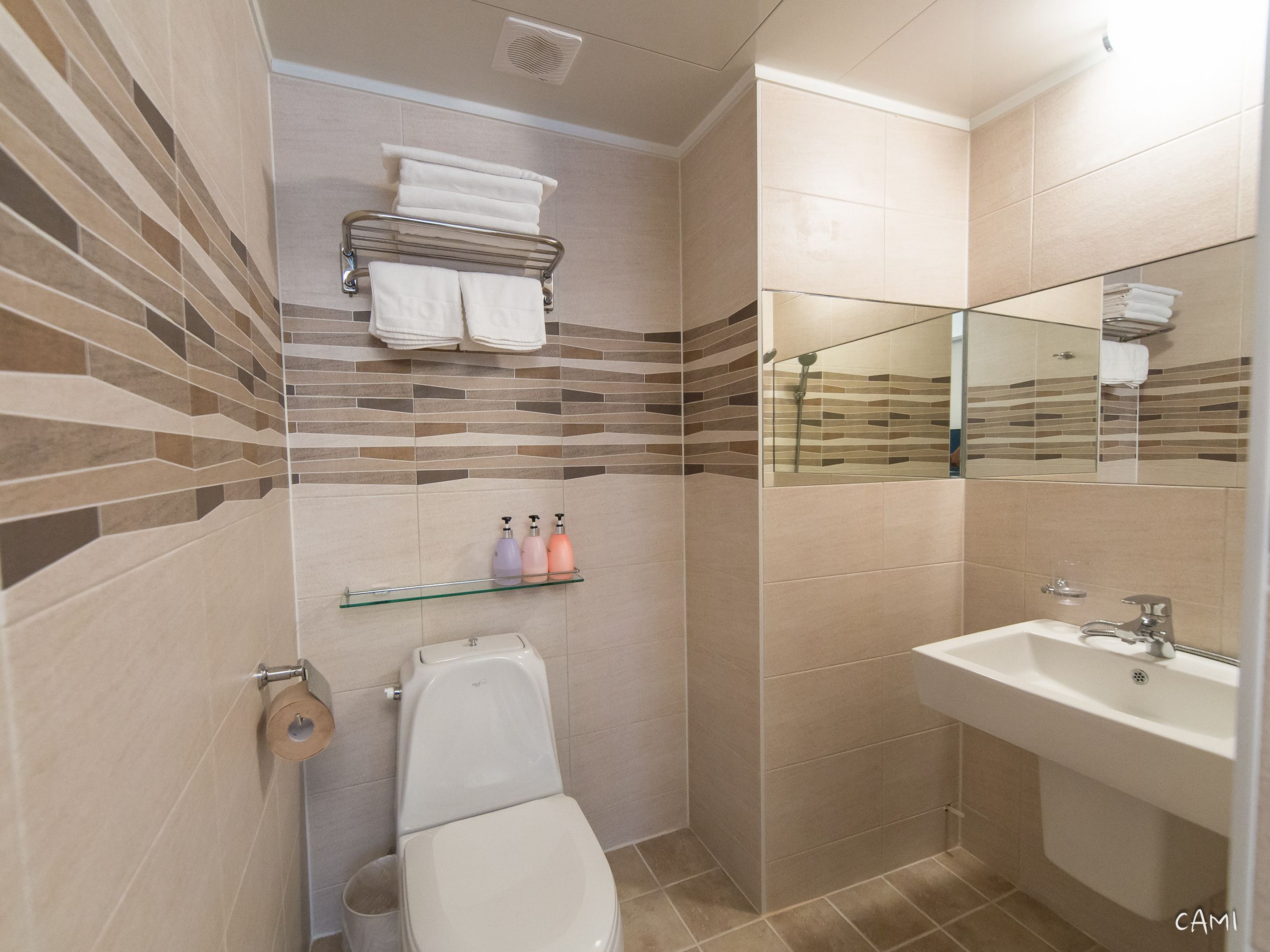 Ein Badezimmer mit Toilette, Waschbecken und Spiegel. Handtücher und Pflegeprodukte liegen auf Regalen.