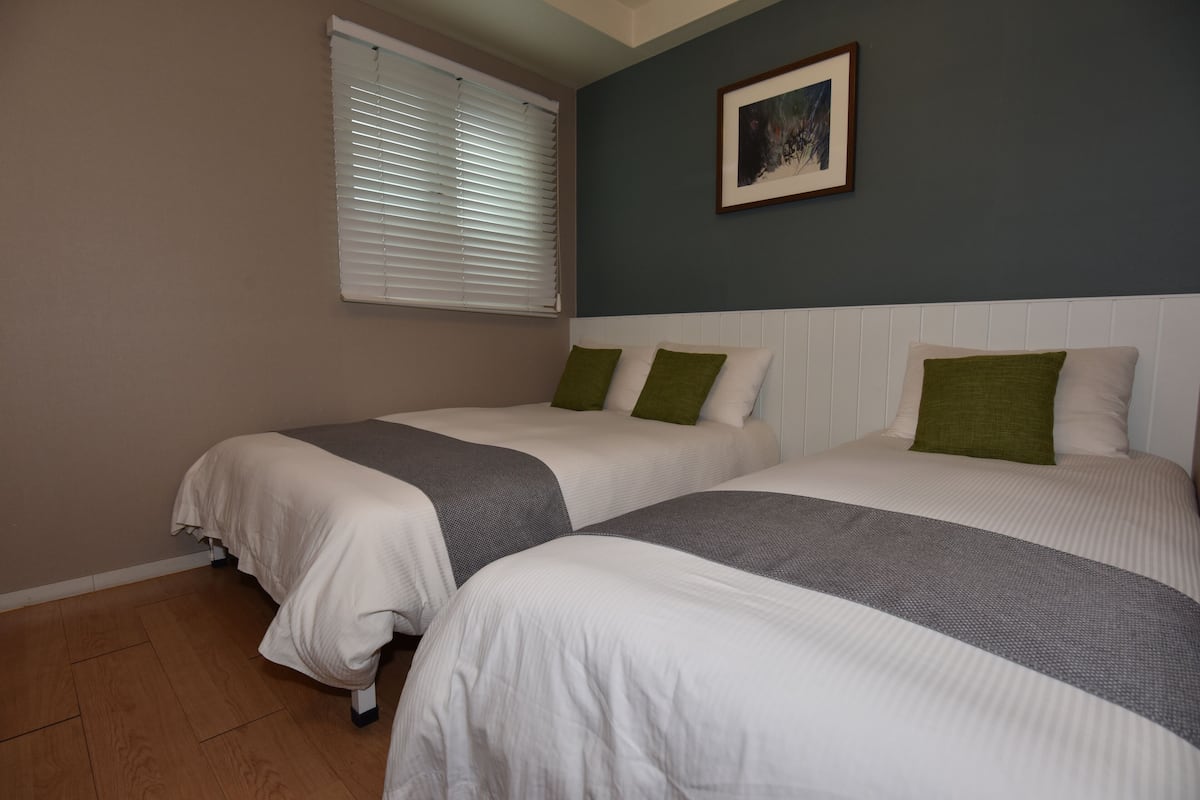 Une chambre avec deux lits, une fenêtre avec des stores, un tableau encadré au mur et un plancher en bois.