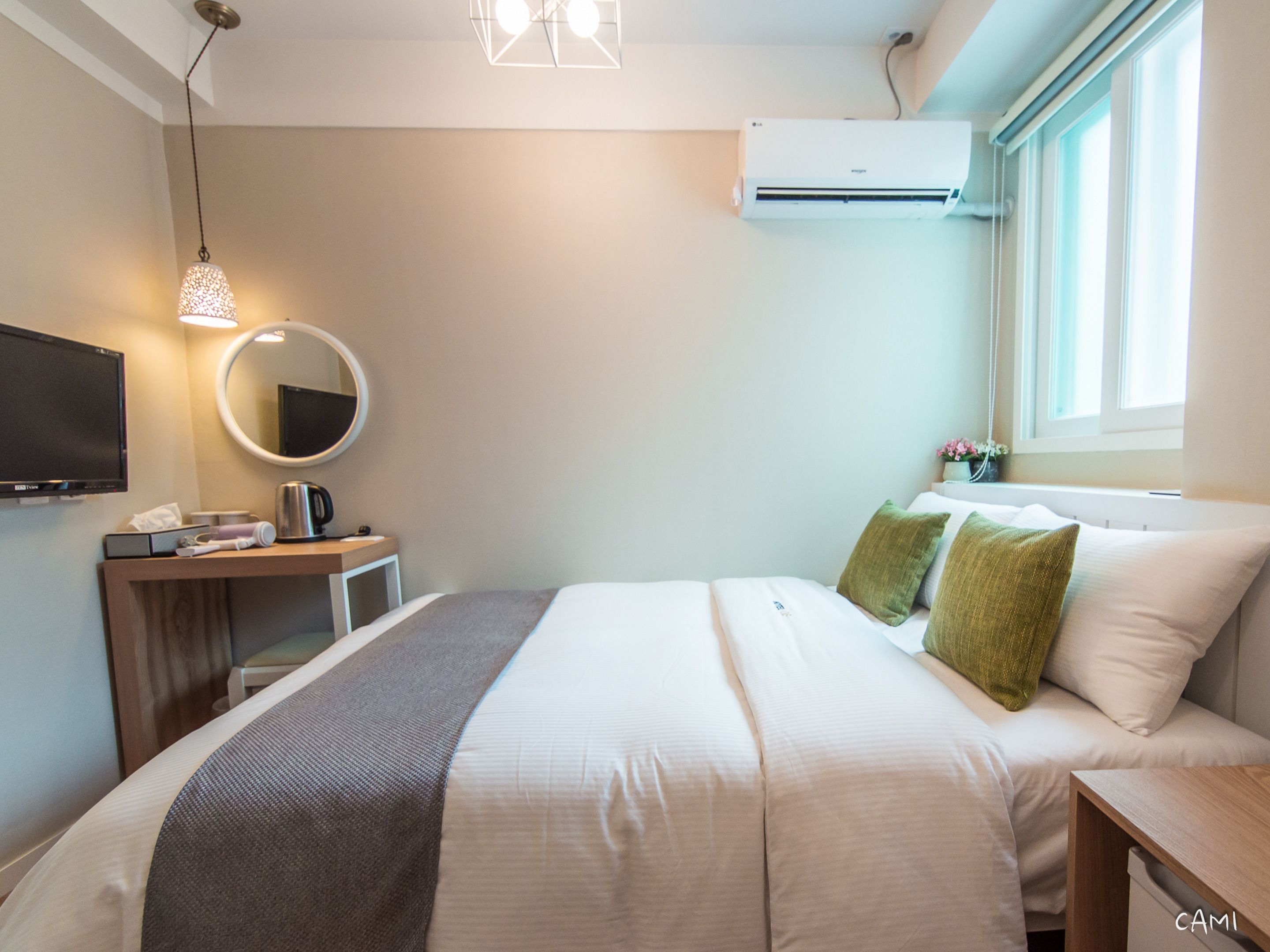 A hotel room with a bed, a desk with a computer, a TV, a mirror, an air conditioner, and a window with a view.