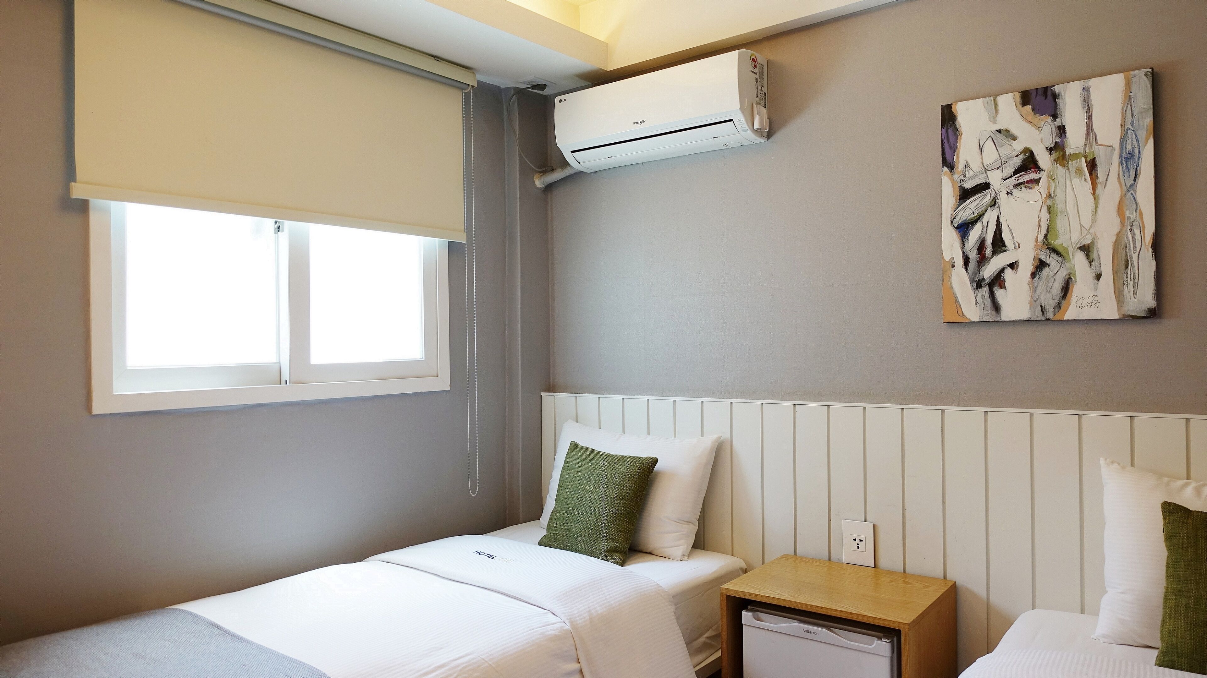 Une chambre d’hôtel avec un lit simple, une table de chevet avec une lampe, un petit meuble de chevet, une fenêtre avec un store, un appareil de climatisation et une œuvre d’art abstraite accrochée au mur.