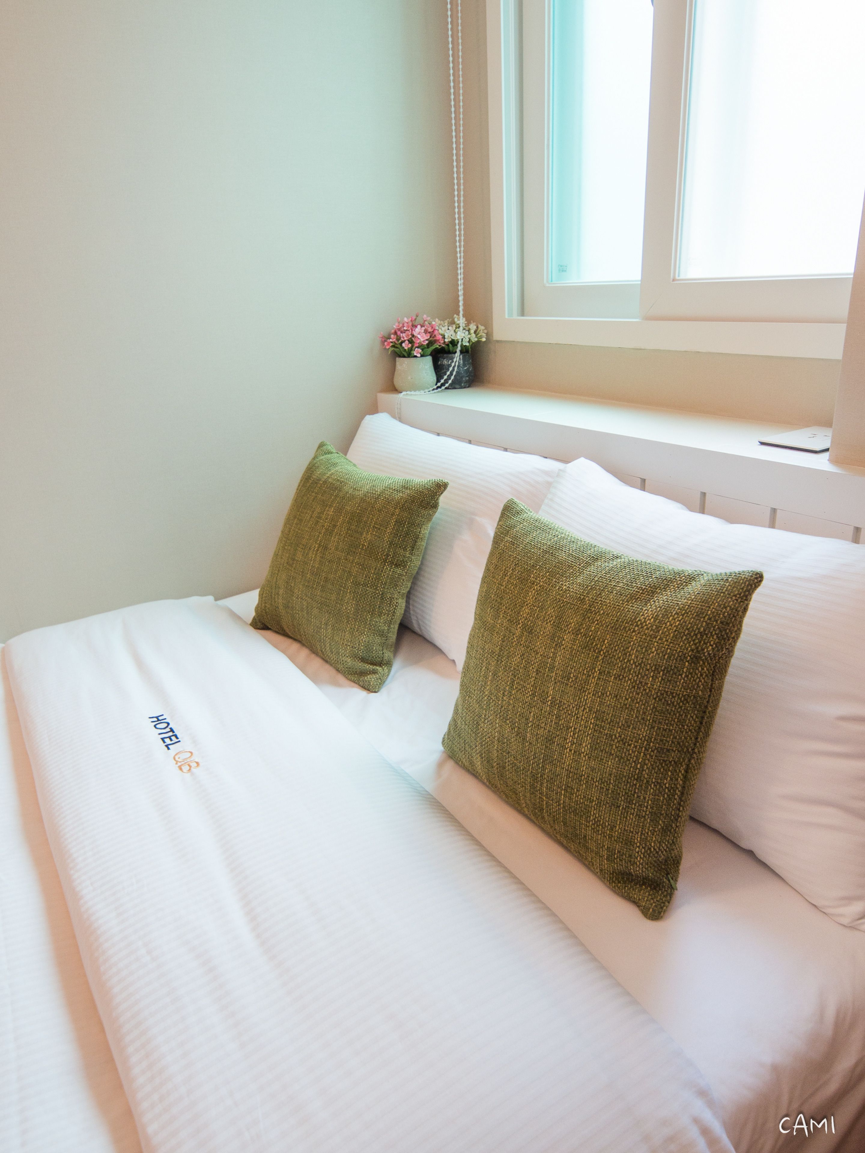A neatly made bed with white linens and two green pillows, a small potted plant with pink flowers on a windowsill, and a white wall.
