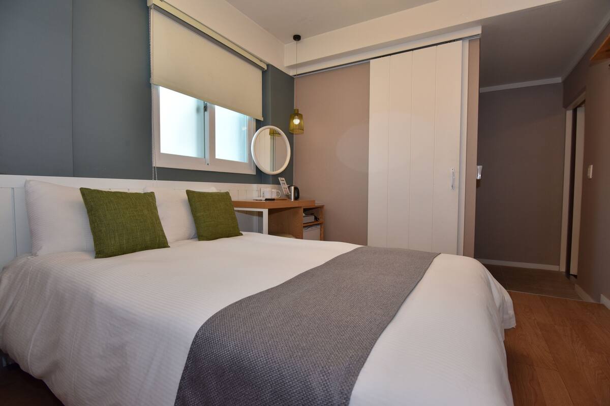 Un lit bien fait, avec une couette blanche et deux oreillers verts, une table de chevet avec une lampe et un miroir au-dessus.