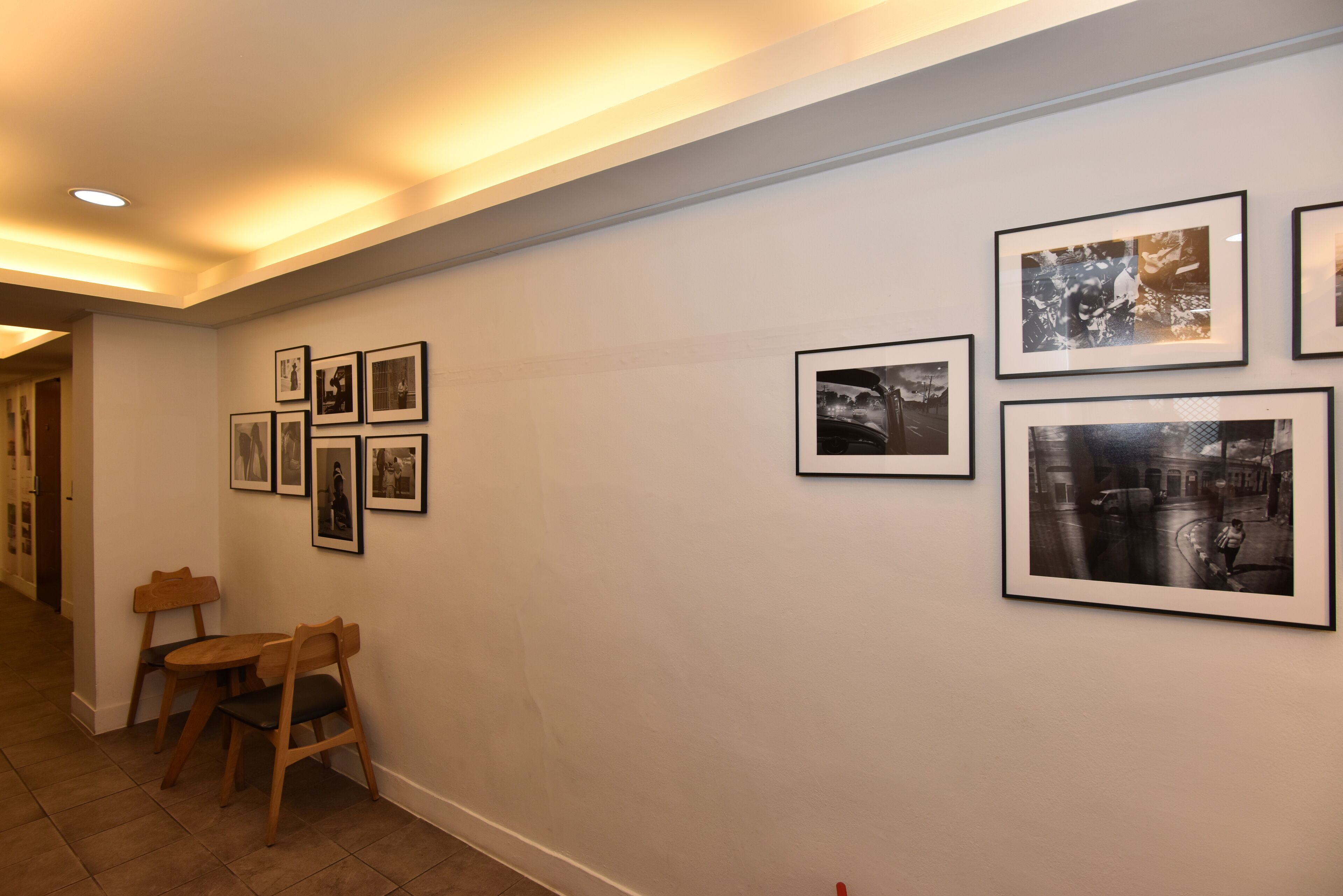 A hallway with framed photographs on the wall, wooden chairs, and a tiled floor.