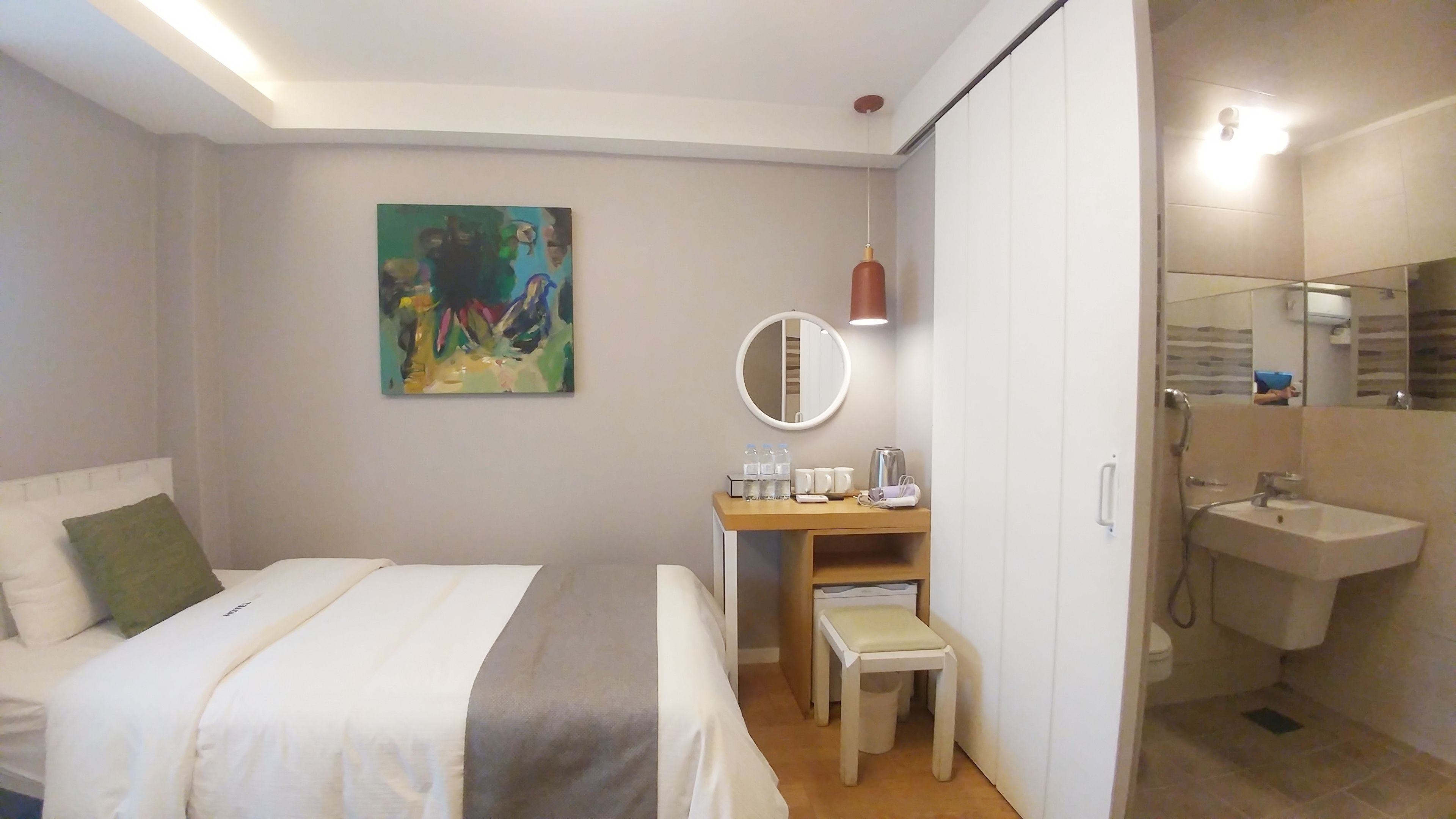 A hotel room with a bed, a desk with a chair, a small table, a mirror, and a bathroom with a sink and mirror.