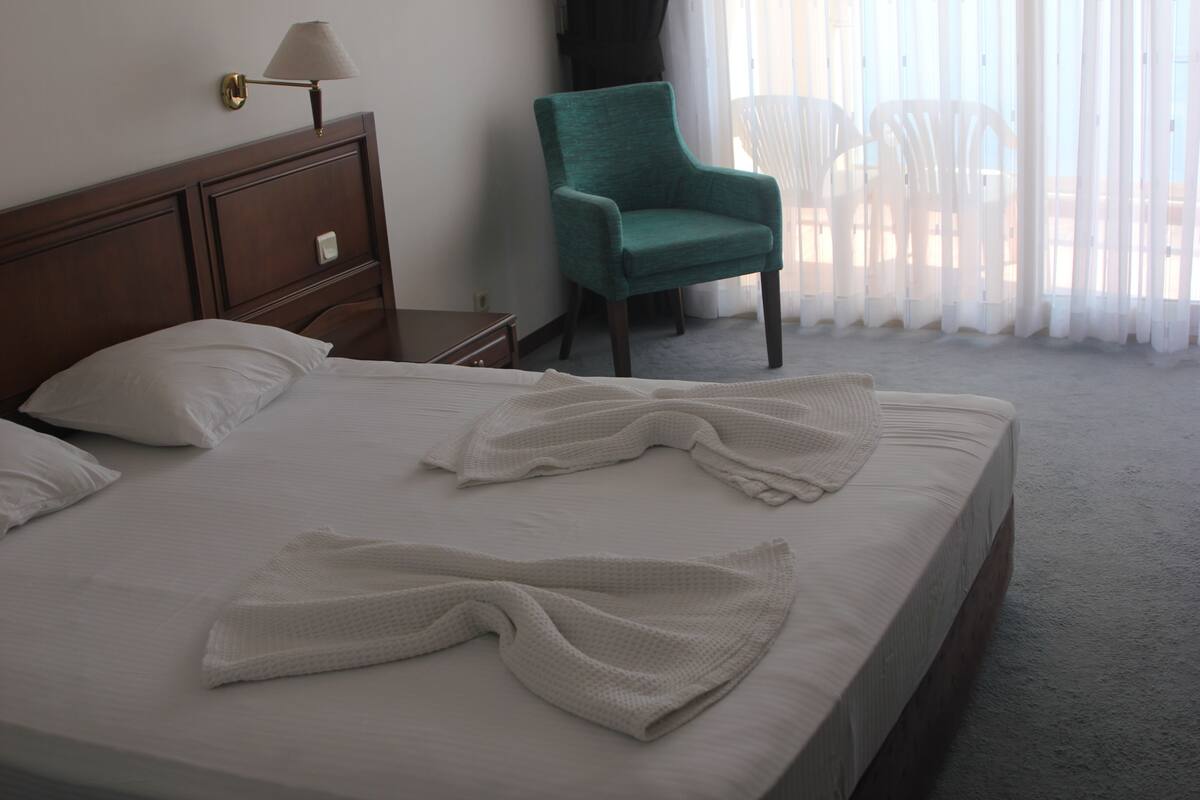 A neatly made bed with white linens and towels, a wooden nightstand with a lamp, a green armchair, and a view of white chairs outside through sheer curtains.
