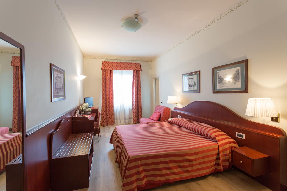 Een hotelkamer met een bed, nachtkastjes, een bureau, een stoel, een spiegel en een raam met gordijnen.