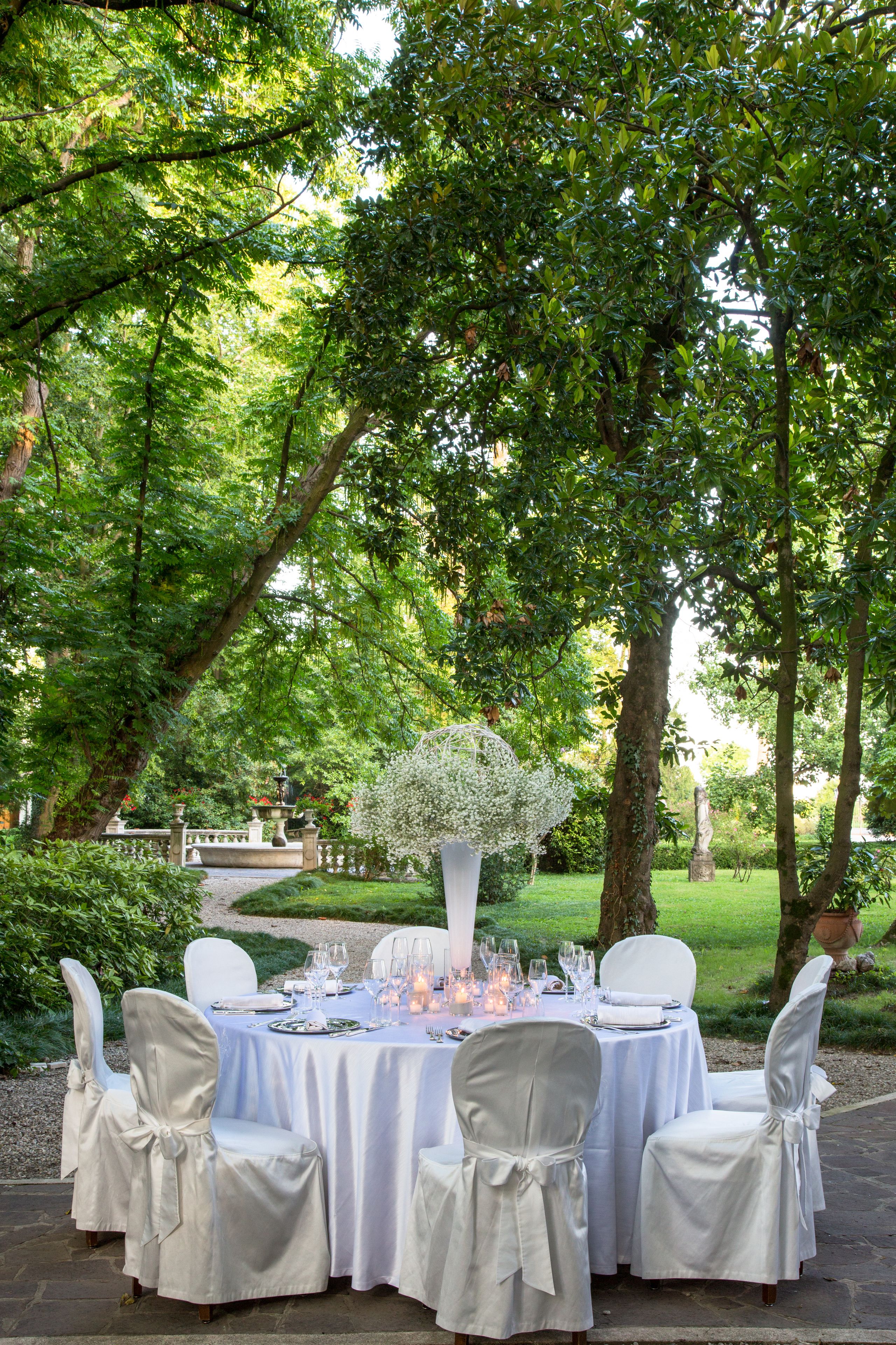 An outdoor dining setup with white tablecloths and chairs, surrounded by lush greenery and trees.