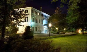 A large, multi-story building with a symmetrical facade, illuminated windows, and a central entrance with a staircase. - Villa Revedin (Gorgo al Monticano)