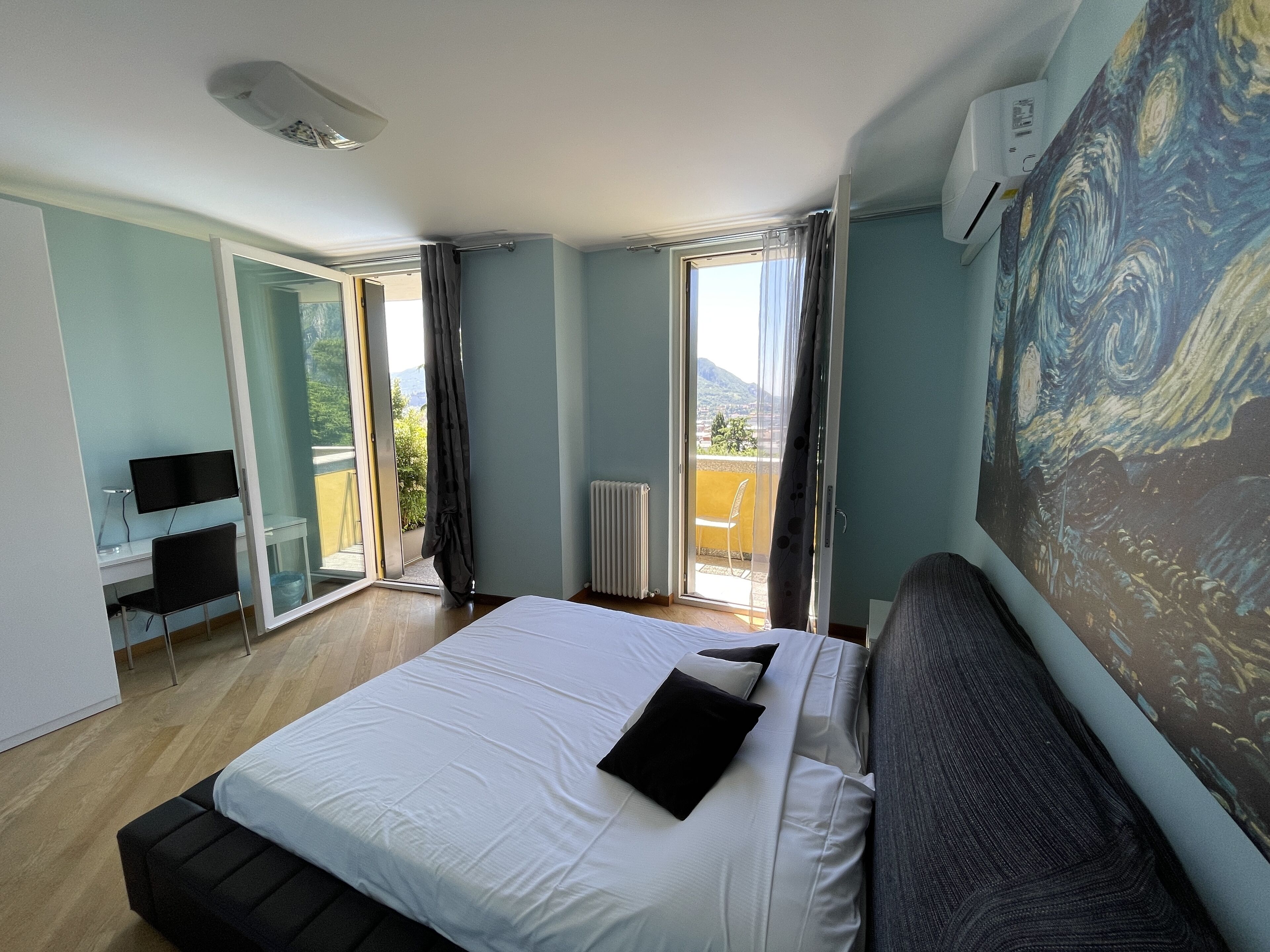 A bedroom with a large bed, a desk with a chair, a painting on the wall, and a balcony with a view.