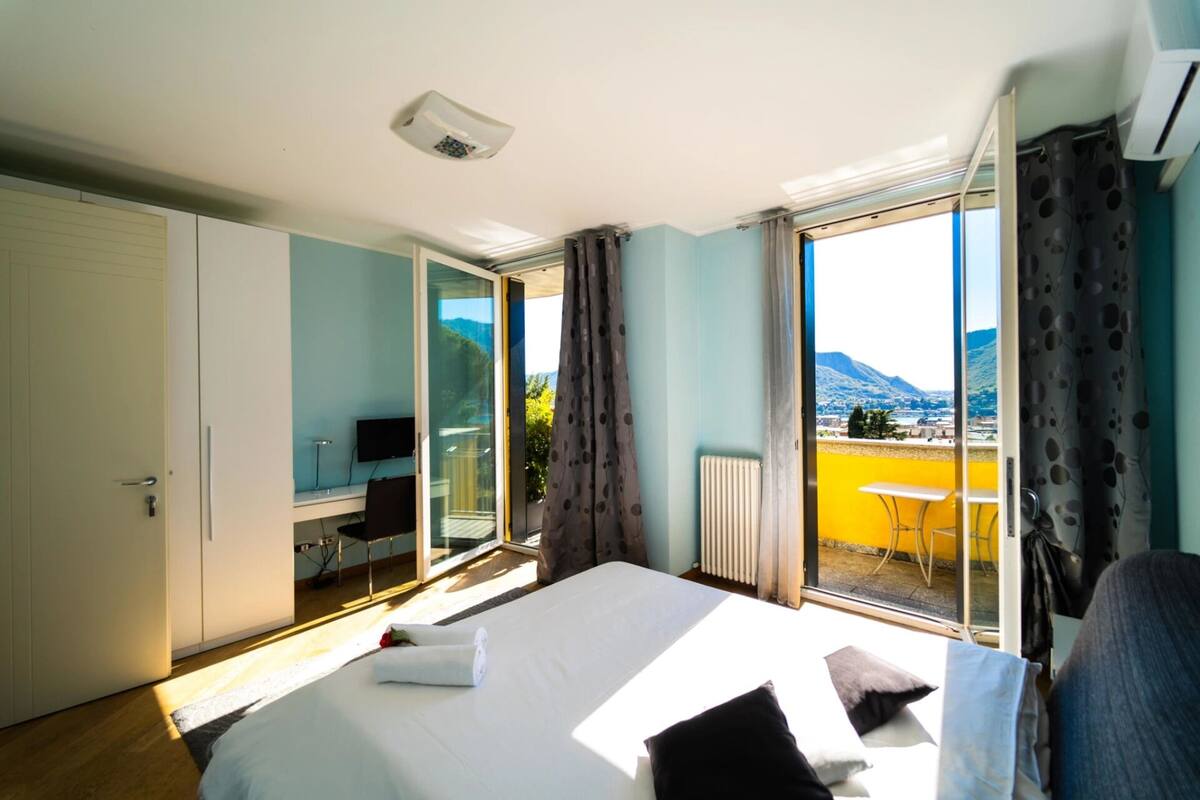 A bedroom with a bed, a desk, and a view of a balcony with a table and chairs.