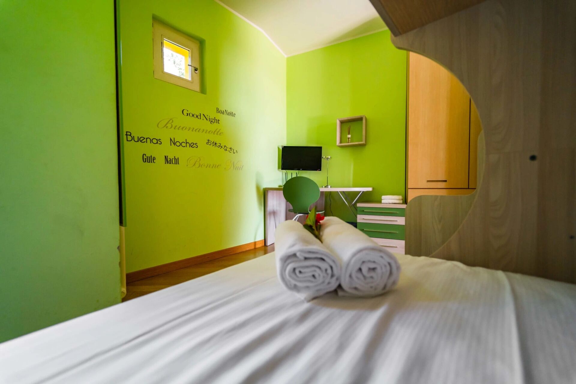 A hotel room with a bed, a desk, a chair, a small window, and a green wall with multilingual 'Good Night' text.