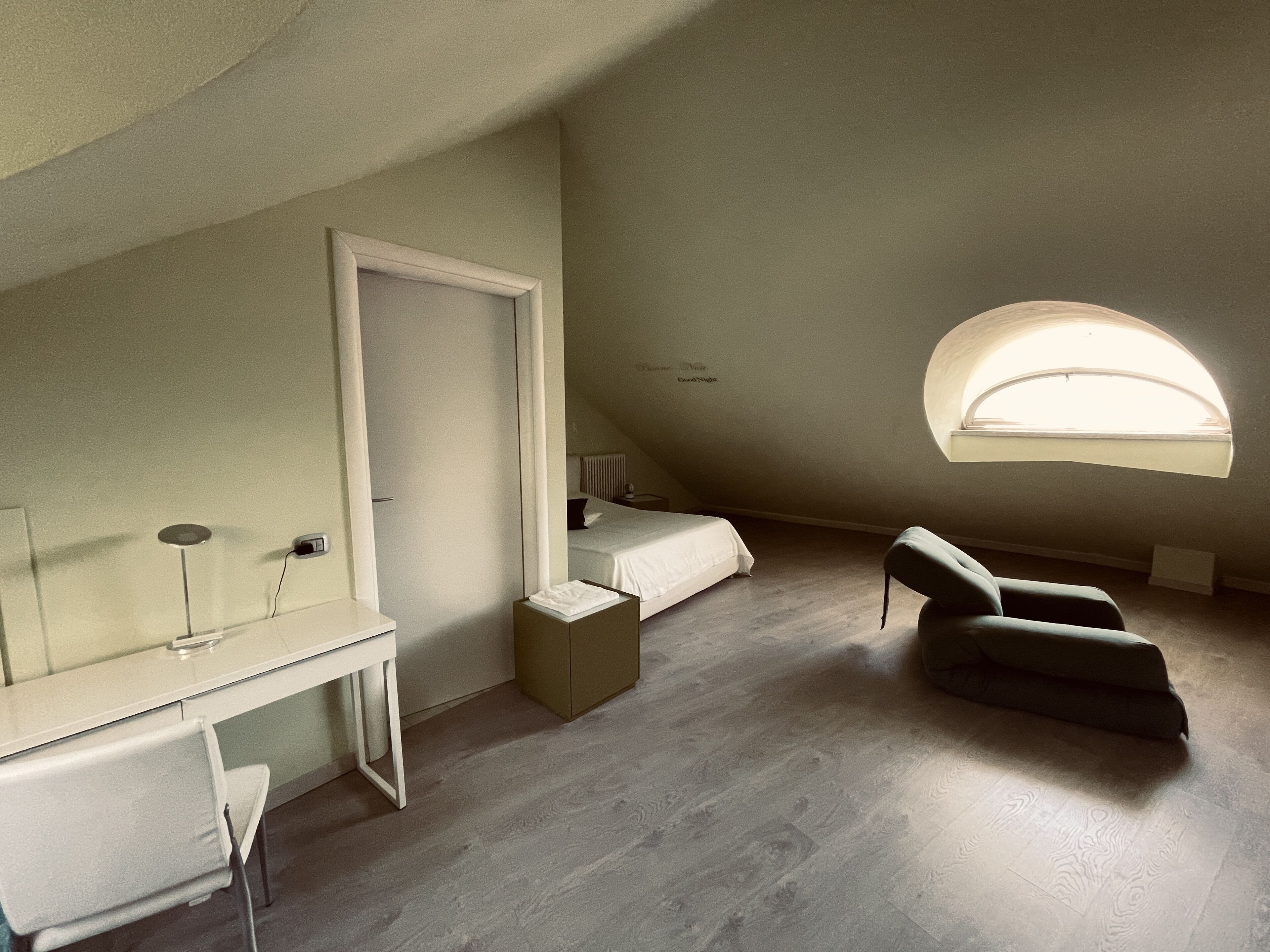 A small, well-lit attic bedroom with a desk, a chair, a bed, and a large window.