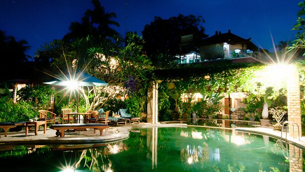 A nighttime pool area with lounge chairs and tables, surrounded by lush greenery and a building in the background. - Kusnadi Hotel (Legian)