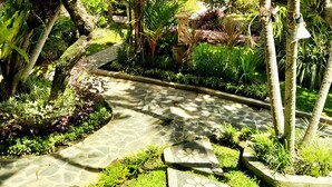 A lush garden with a stone pathway, various tropical plants, and a building with a tiled roof in the background. - Kusnadi Hotel (Legian)