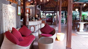 A cozy seating area with wicker furniture and red cushions, a stone wall with carvings, and a bar area with wooden decor and plants. - Kusnadi Hotel (Legian)