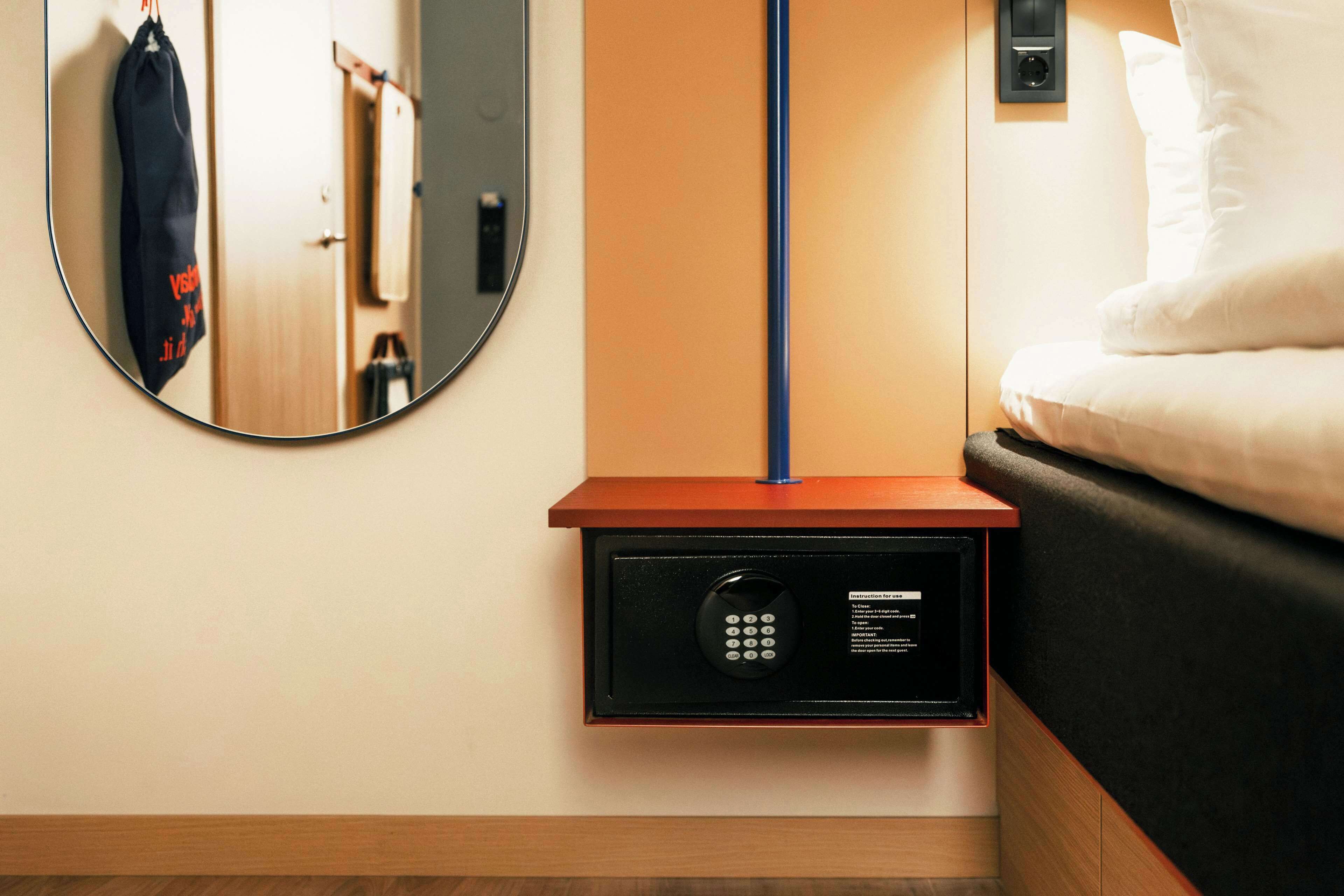A hotel room with a bedside safe, a round mirror, a wooden nightstand, and a bed with white bedding.