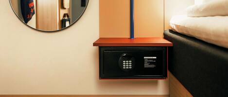 A hotel room with a bedside safe, a round mirror, a wooden nightstand, and a bed with white bedding.