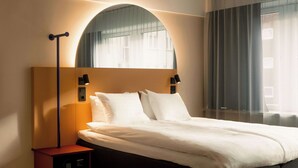 Una habitación de hotel moderna con una cama grande, una mesita de noche con lámpara y un ventanal con cortinas.