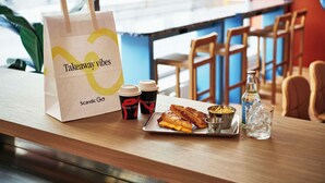 Una bolsa para llevar, dos vasos, un sándwich, un cuenco pequeño y una botella de agua sobre una mesa de madera en un café.