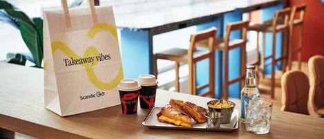 A takeaway bag, two cups, a sandwich, a small bowl, and a bottle of water on a wooden table in a cafe setting.