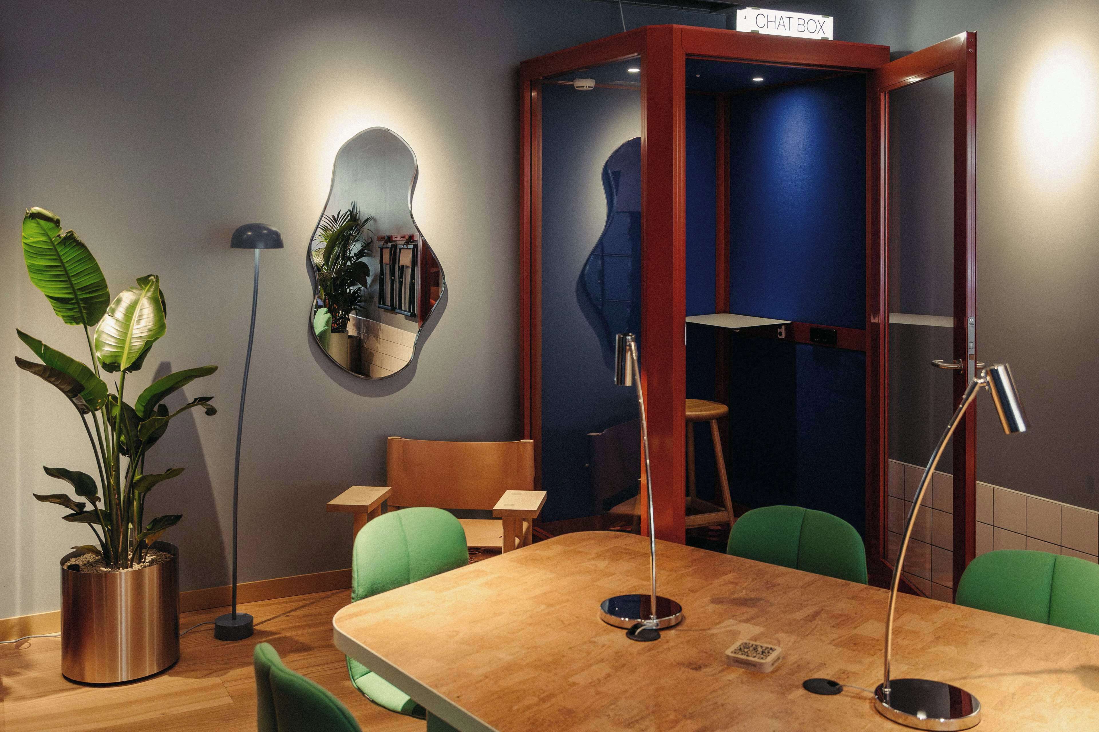 A conference room with a wooden table, green chairs, a potted plant, a mirror, and a 'CHAT BOX' sign.