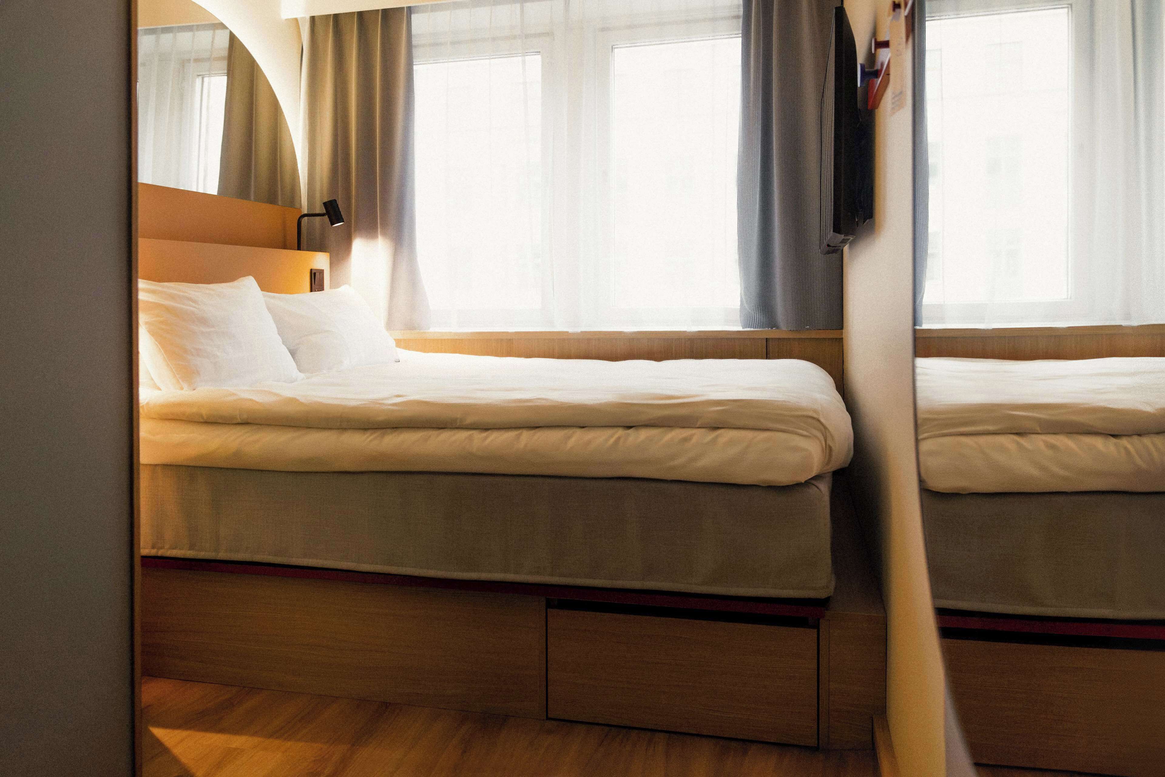 A hotel room with two beds, a wooden headboard, a bedside table, and a window with curtains.