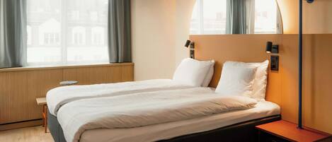 Habitación de hotel con cama doble, mesita de noche, lámpara y un amplio ventanal con cortinas.