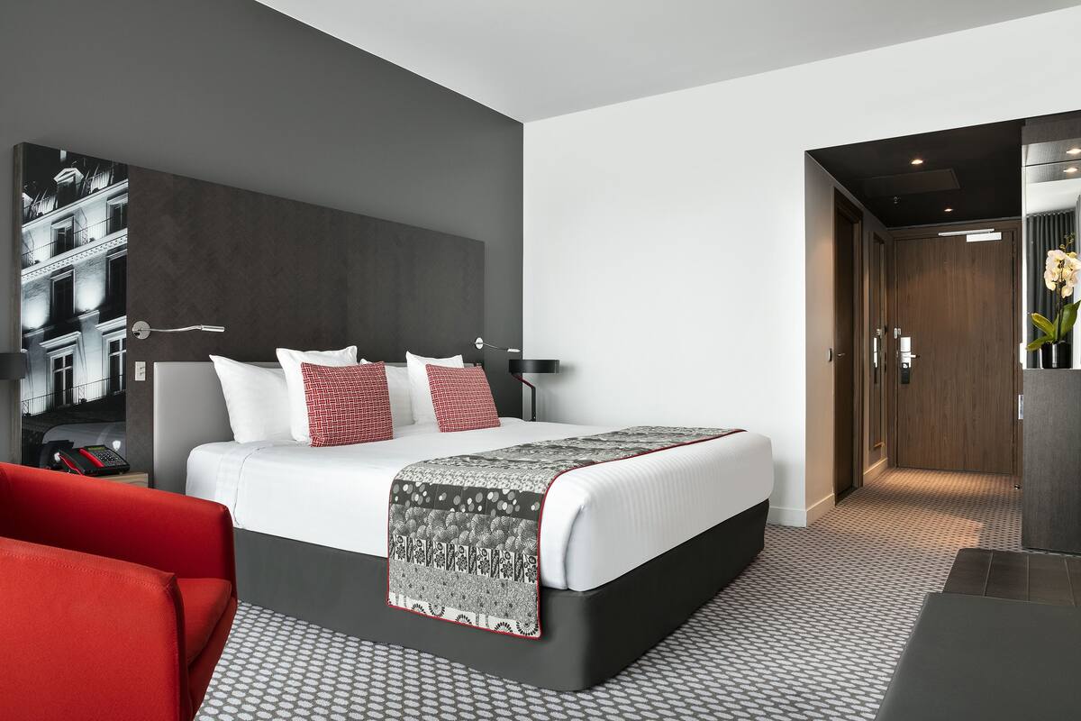 A modern hotel room with a large bed, a red chair, a patterned throw, and a view of the city through the window.
