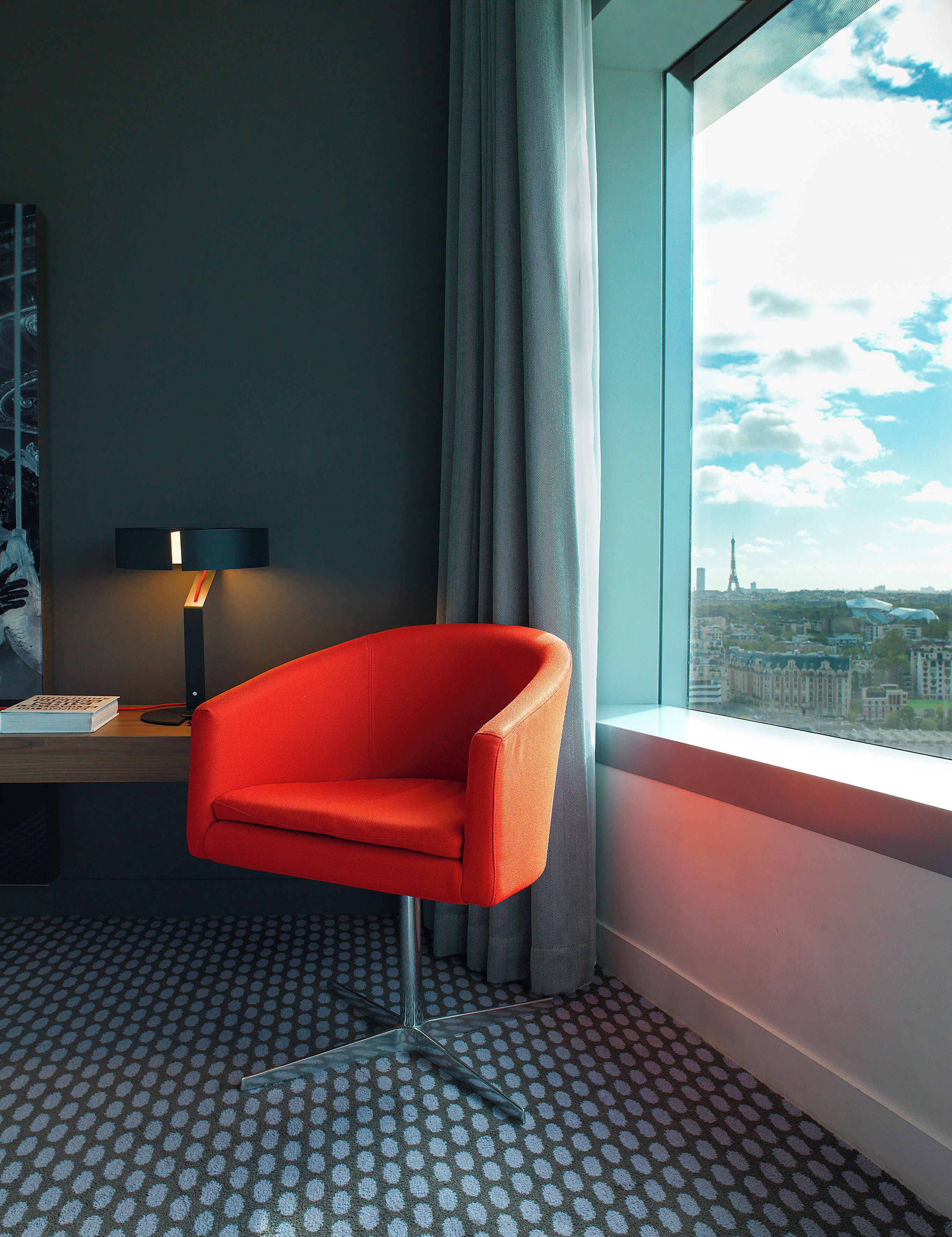 A hotel room with a red armchair, a desk with a lamp, and a view of a cityscape through a window.
