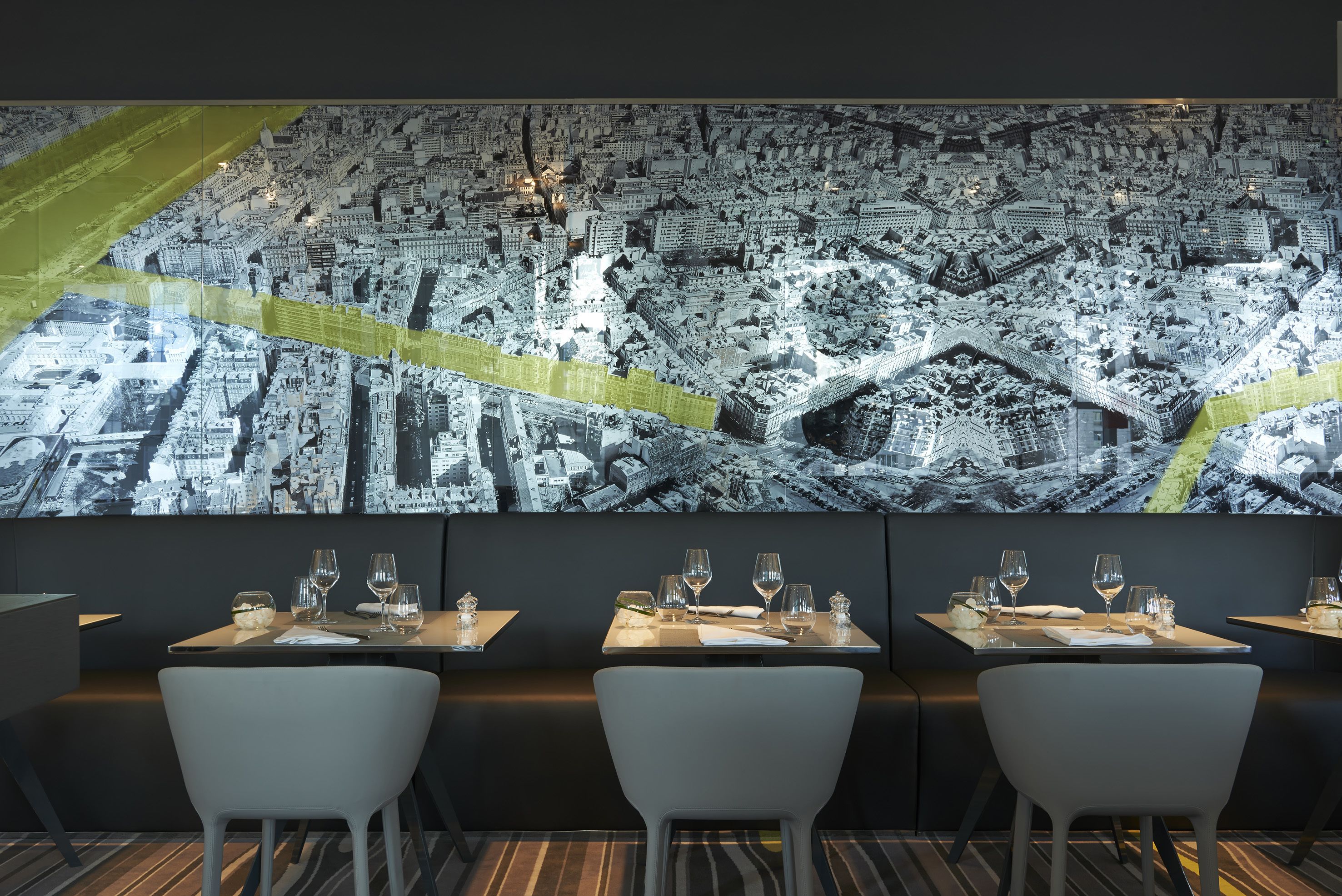 A restaurant with a large cityscape mural on the wall, tables set with wine glasses and plates, and modern white chairs.