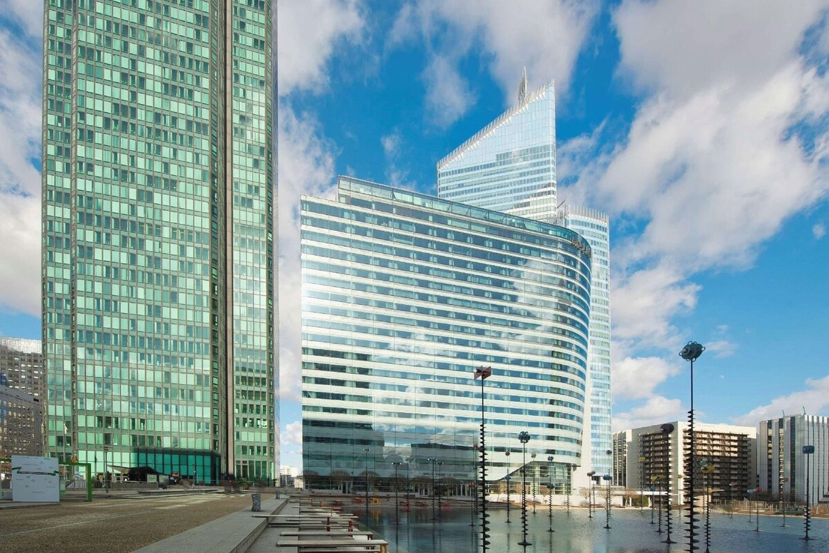 A modern building with a curved facade and a distinctive spire, surrounded by a water feature and street lamps.