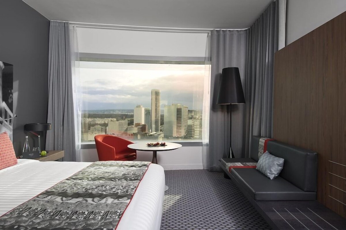A hotel room with a large bed, a red chair, a small table with fruit, a sofa, and a view of a cityscape through large windows.