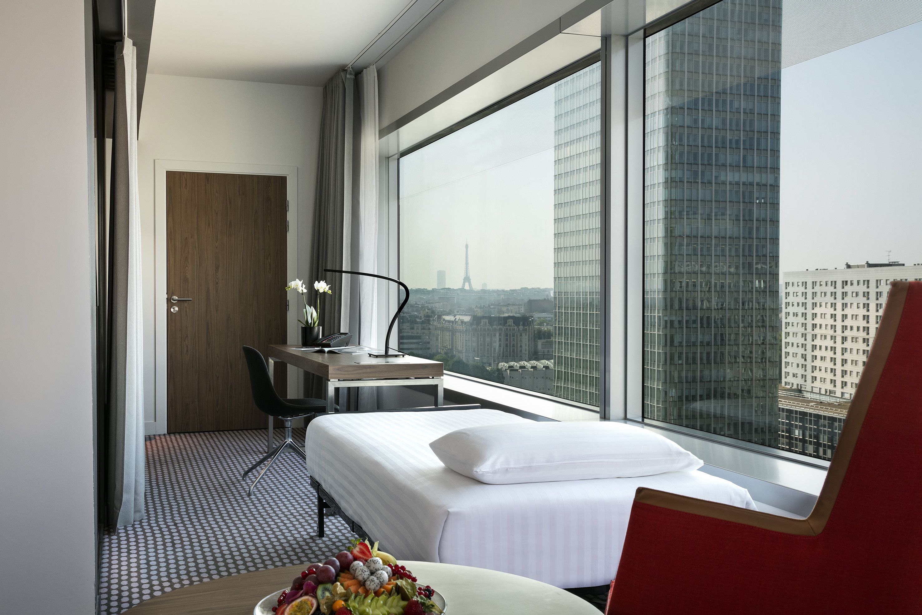 Ein Hotelzimmer mit einem Bett, einem Schreibtisch, einem Sessel, einem Esstisch mit Früchten und einem großen Fenster mit Blick auf die Stadt.