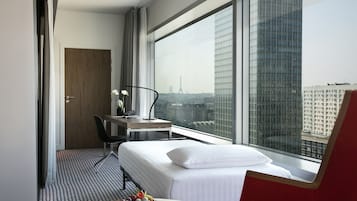 Ein Hotelzimmer mit einem Bett, einem Schreibtisch, einem Sessel, einem Esstisch mit Früchten und einem großen Fenster mit Blick auf die Stadt.
