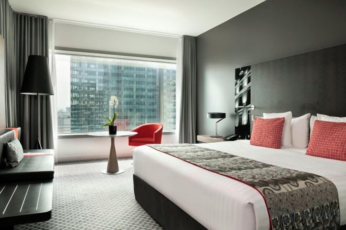 A modern hotel room with a large bed, a desk, a red chair, and a view of the city.