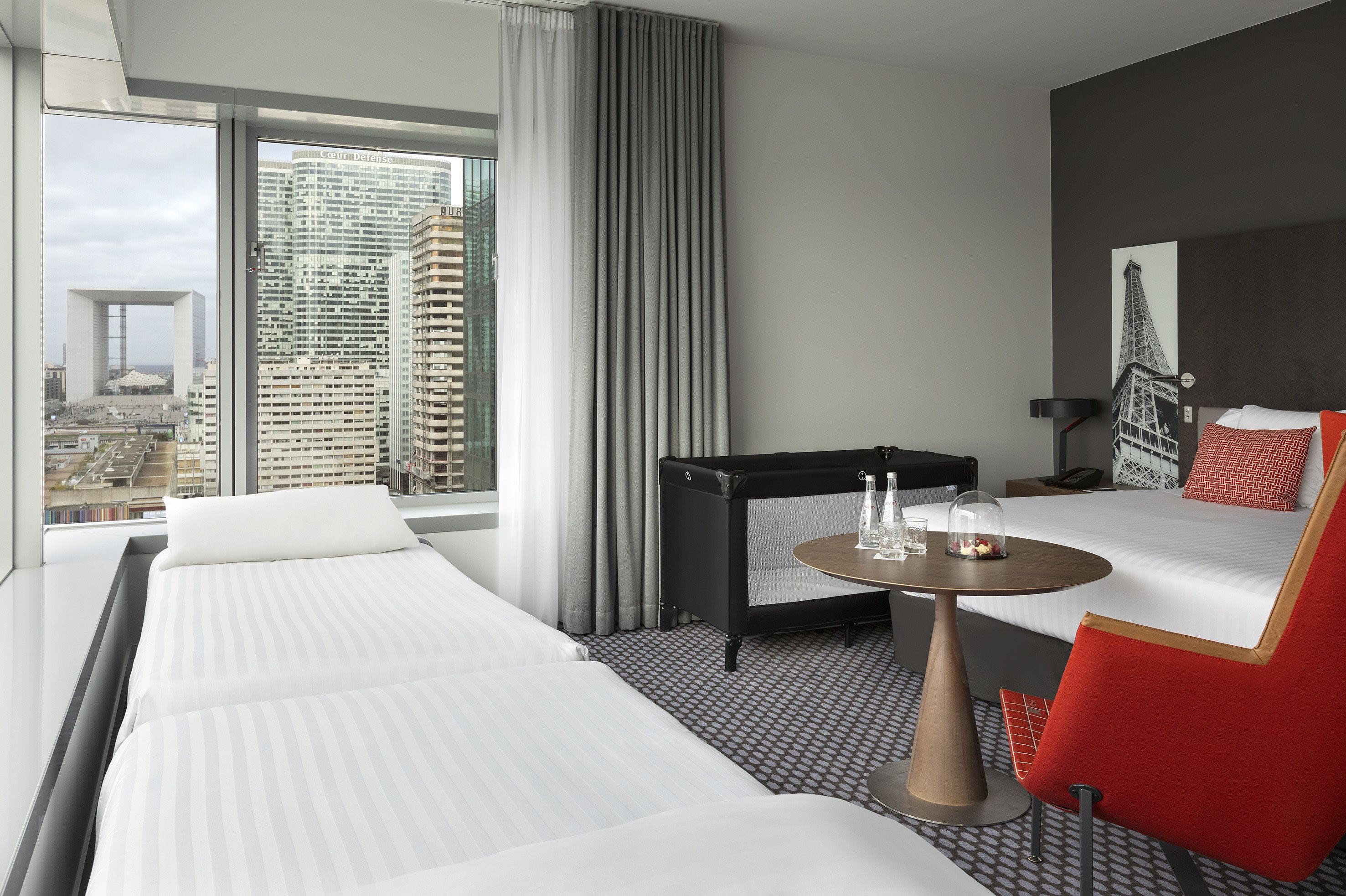 A hotel room with a large window, a bed, a desk, a chair, and a view of a cityscape.