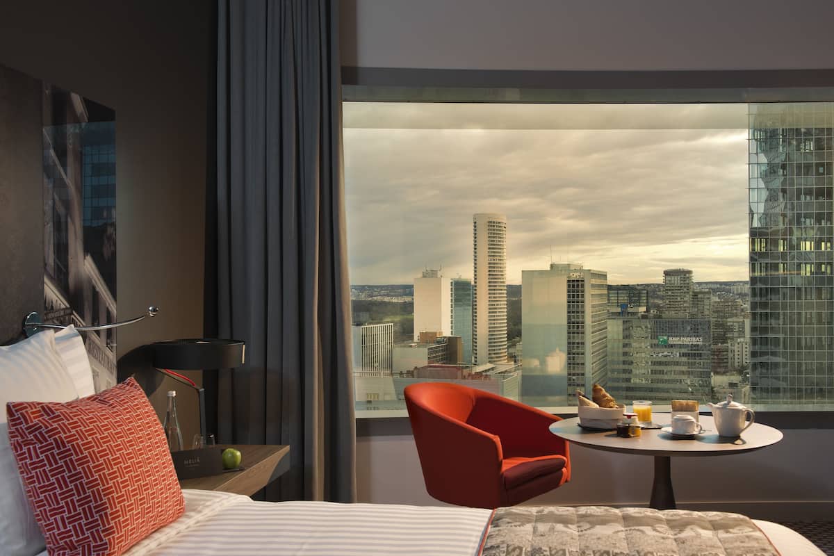 A hotel room with a bed, a red chair, a small table with breakfast items, and a large window offering a city view.