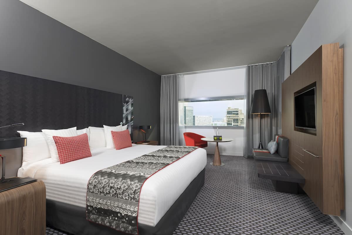A modern hotel room with a large bed, a TV, a desk, and a seating area by the window.