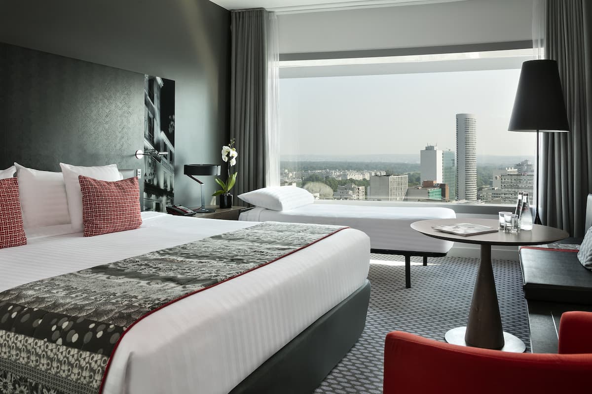 A modern hotel room with a large bed, a sofa, a small table, and a view of the cityscape.