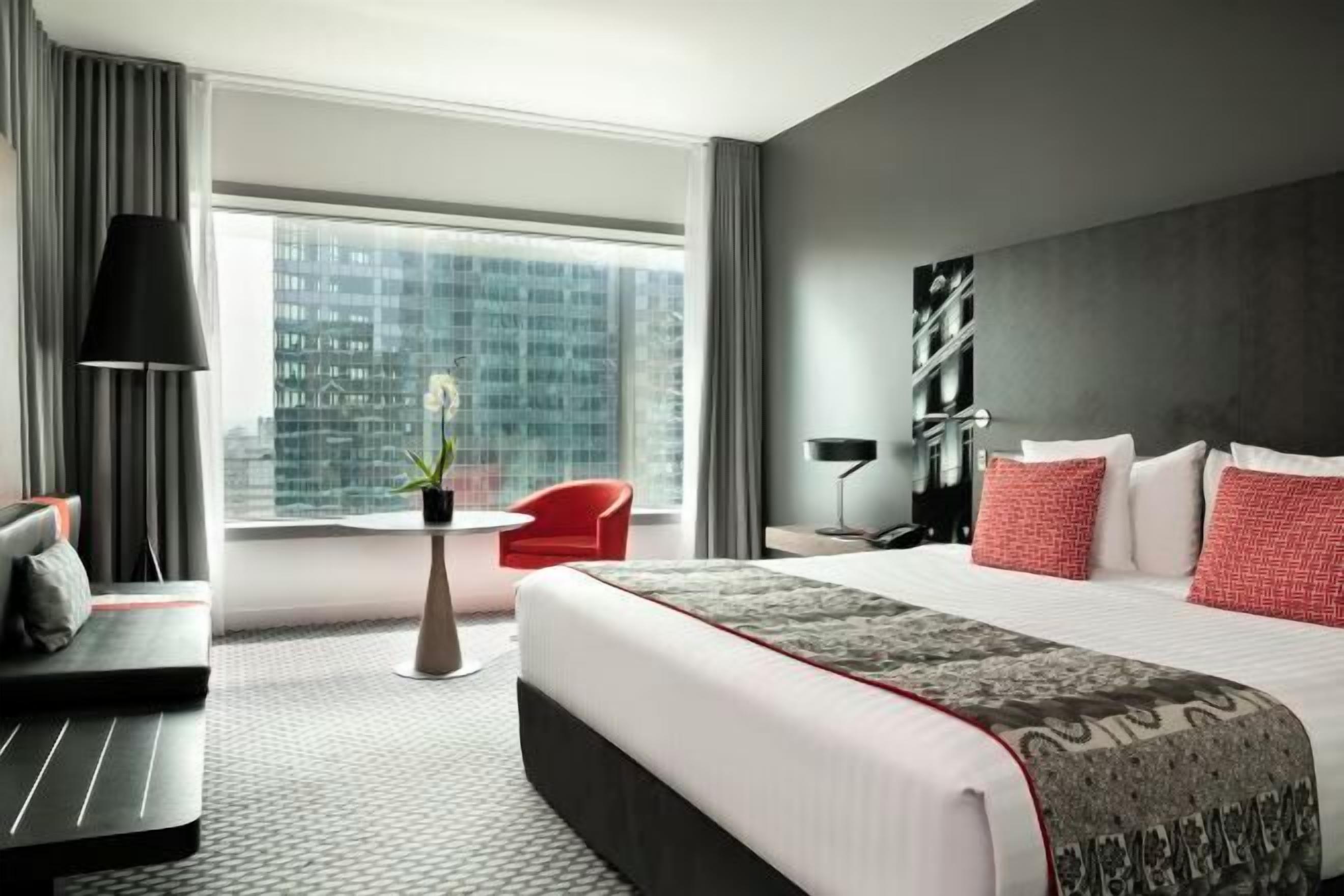 A modern hotel room with a large bed, a red chair, a small table with a plant, and a city view through the window.