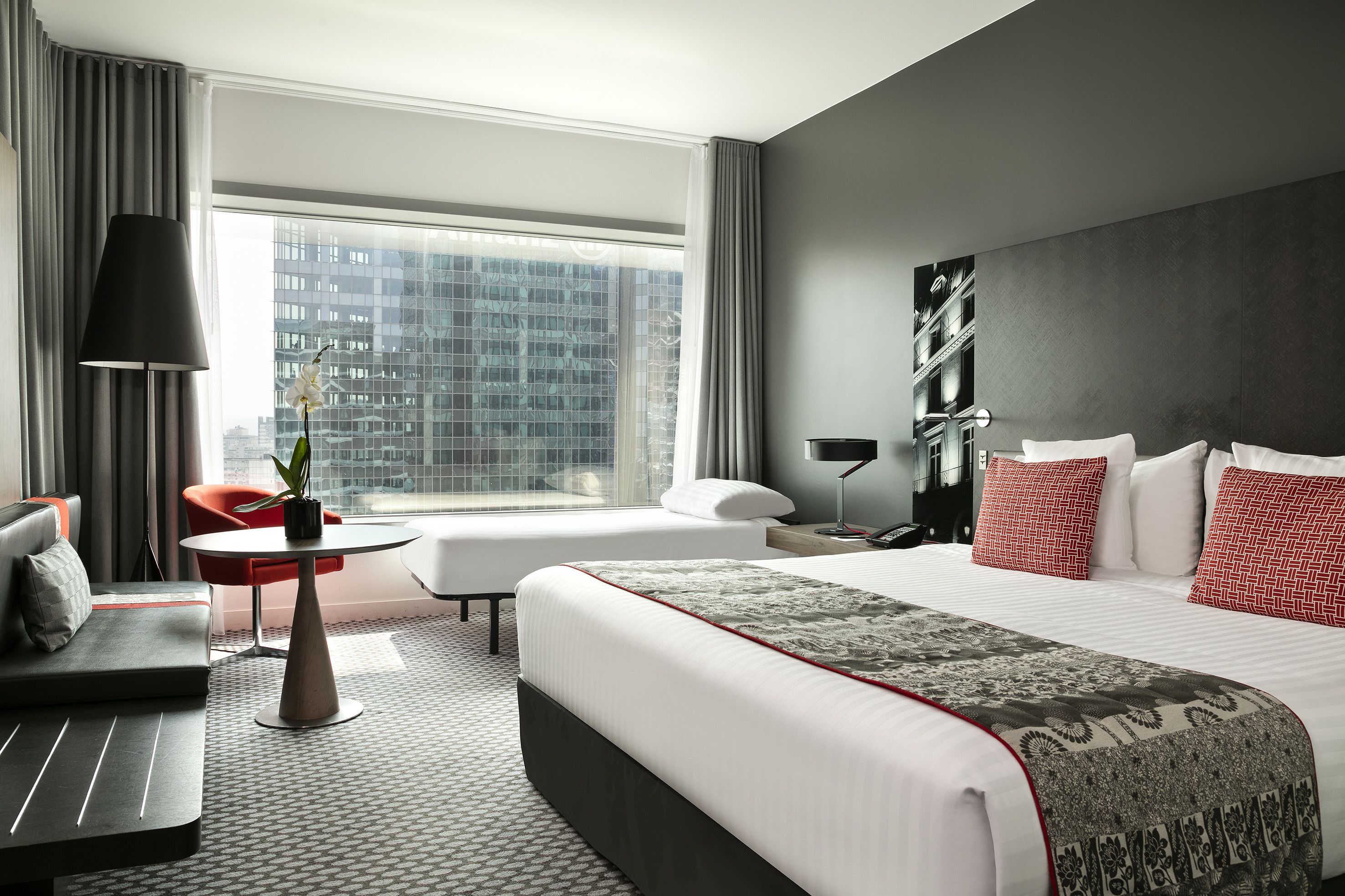 A modern hotel room with a large bed, a sofa, a small table, and a view of a cityscape through large windows.