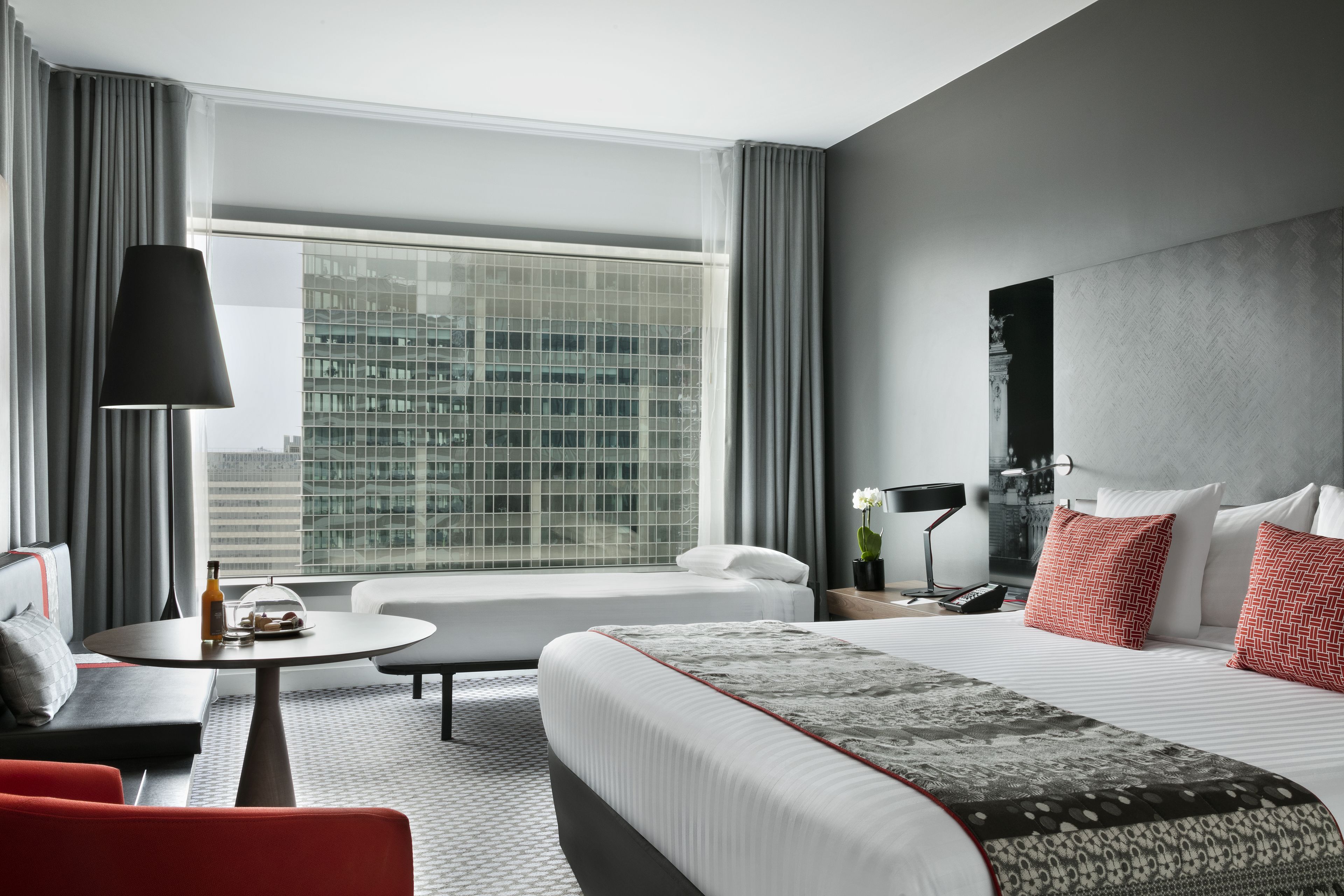A modern hotel room with a large bed, a sofa, a round coffee table, and a view of a cityscape through large windows.