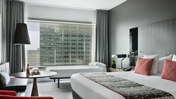 A modern hotel room with a large bed, a sofa, a round coffee table, and a view of a cityscape through large windows.