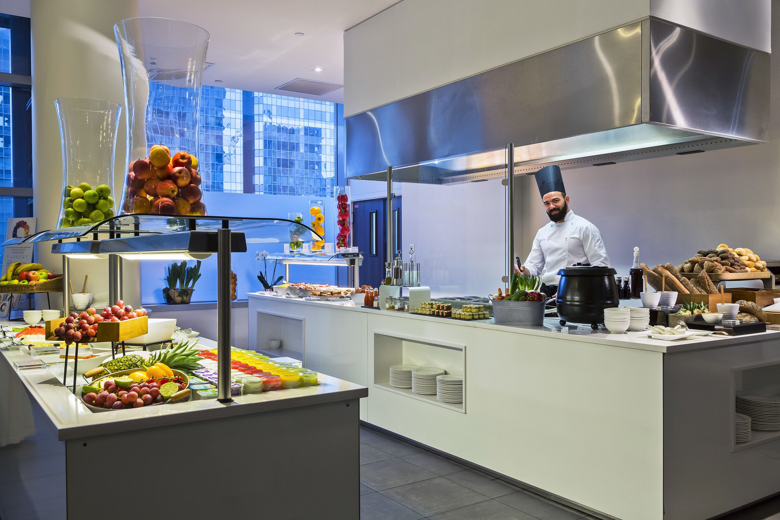 Eine moderne Küche mit einem Koch, der zubereitet, ein Buffet mit Früchten und Gemüse sowie ein Blick auf die Stadt durch große Fenster.