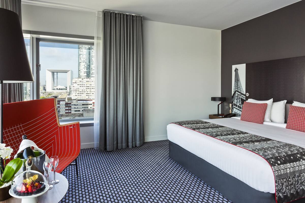 A modern hotel room with a large bed, a city view, a red accent chair, and a dining area with a fruit bowl and glass.