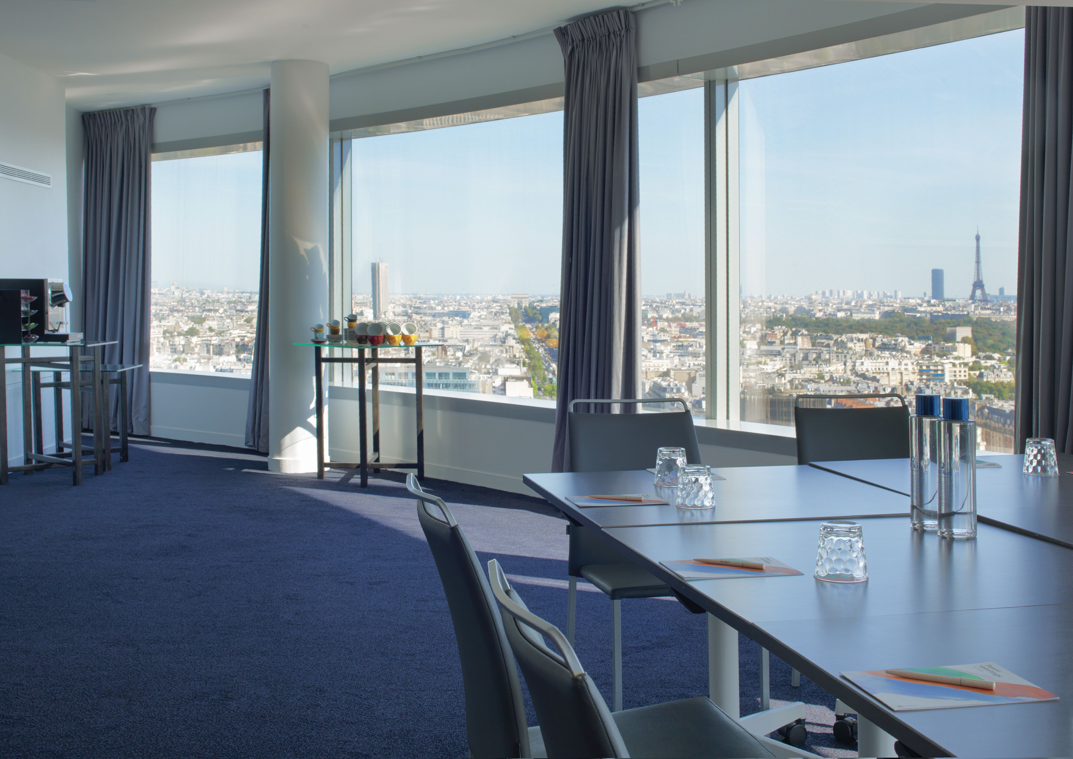 A conference room with a large table, chairs, a small table with cups, and a view of a cityscape through large windows.