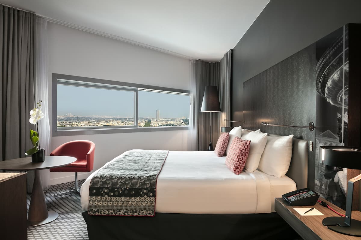 A hotel room with a large bed, a desk, a chair, a window with a city view, and a modern lamp.