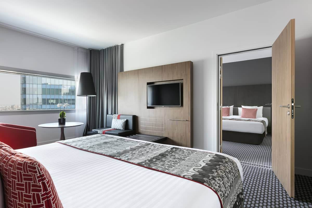 A modern hotel room with a large bed, a TV mounted on the wall, a desk with a chair, and a window with curtains.